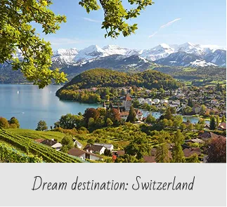 Scenic view of a Swiss village by a lake with green hills and snow-capped mountains in the background.